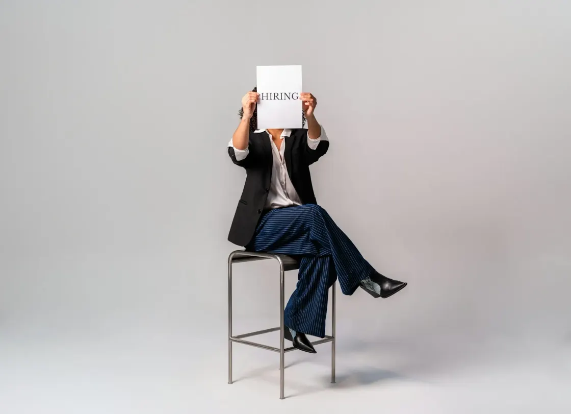 A professional seated on a stool holds a plain white sign reading "HIRING", representing the gap between recruitment signalling and the deeper strategic discipline of employer branding.