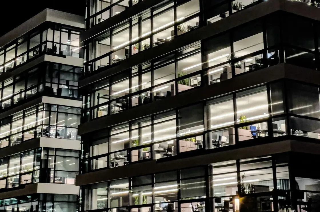 Corporate office building at night, every floor lit, rows of empty desks visible through floor-to-ceiling glass windows.