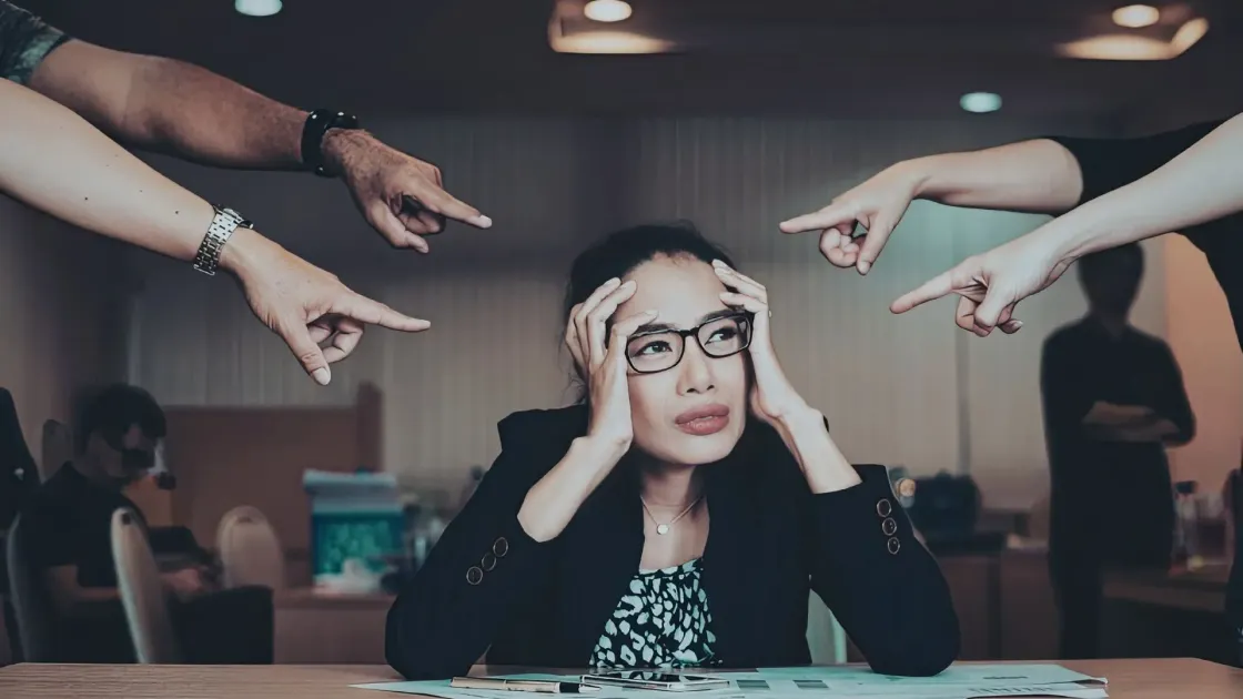 An overwhelmed HR professional holding their head while multiple hands point at them, suggesting workplace pressure, blame, and organisational stress.