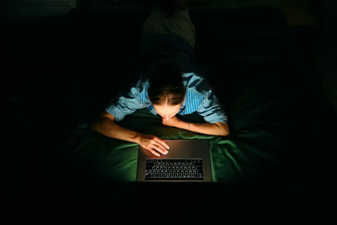 Overhead view of a woman lying in a dark room, face and hands lit only by her laptop screen as she researches late at night.