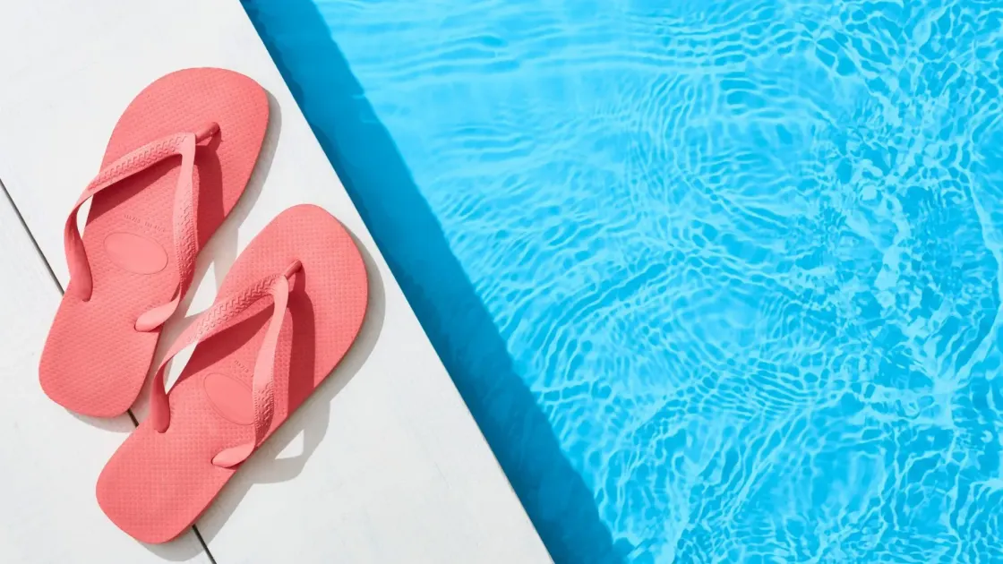 A pair of pink flip-flops resting on a white poolside deck beside bright turquoise water, playing on the idiom "flip-flop" to represent Big Tech reversing its stance on AI-driven layoffs.