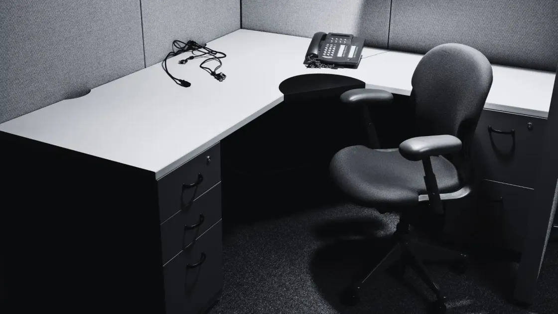 An empty office cubicle in black and white, chair pushed in and a desk phone unplugged with its cables coiled loose, symbolising a role removed by decision rather than by a person.