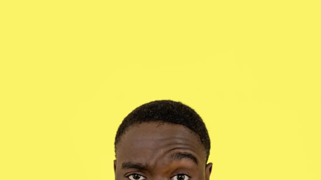 Cropped close on a man's raised eyebrow and narrowed eye, a sceptical look, against a flat yellow background.