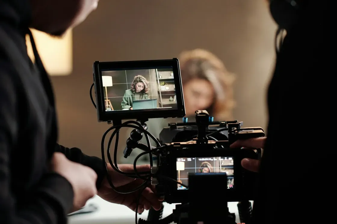 A production crew films an employee seated at a desk, with her image framed on a camera monitor.