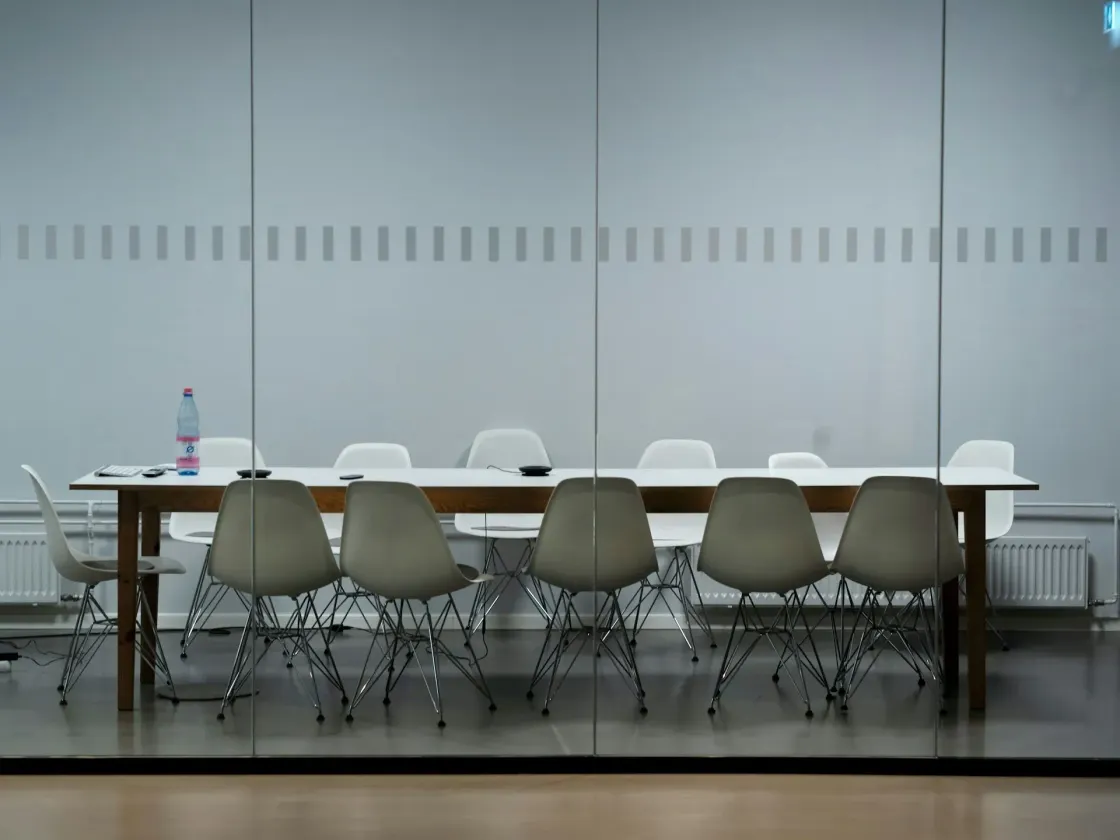 Empty modern conference room with a long meeting table and white chairs behind glass walls.