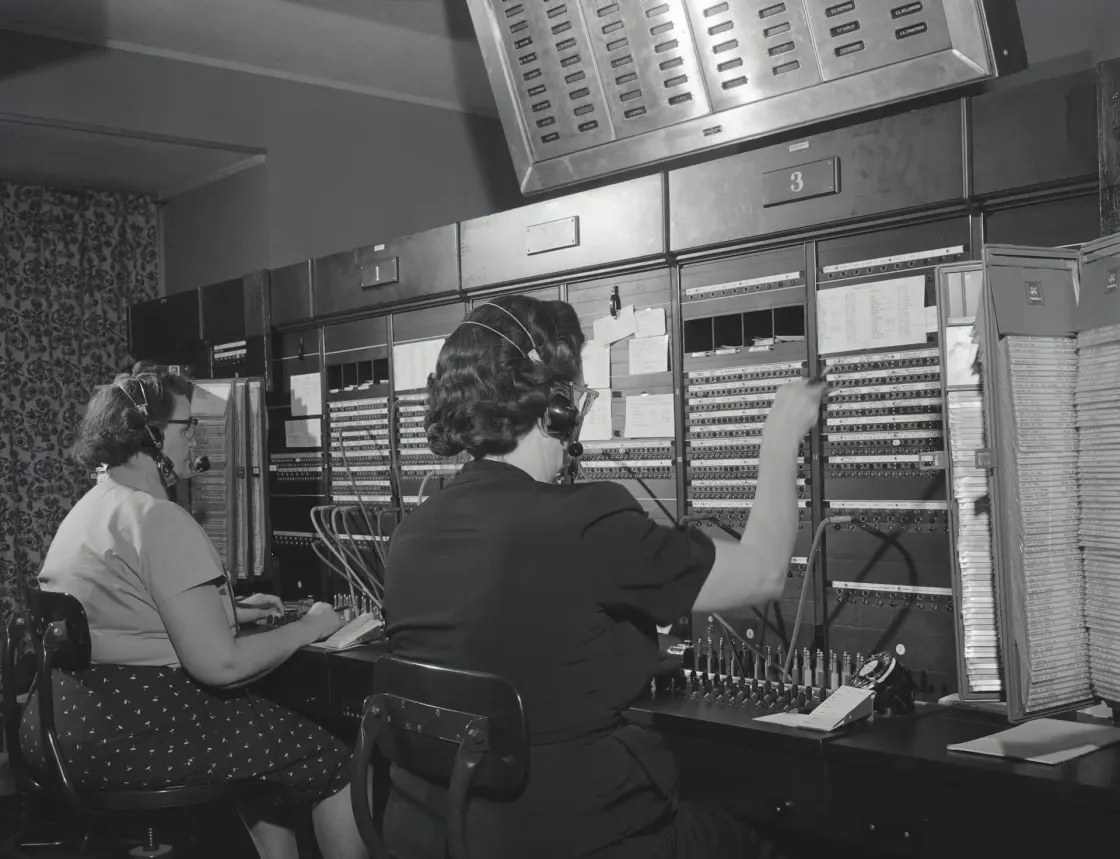 Two telephone operators wearing headsets and connecting calls at a large manual switchboard in a black-and-white photograph.