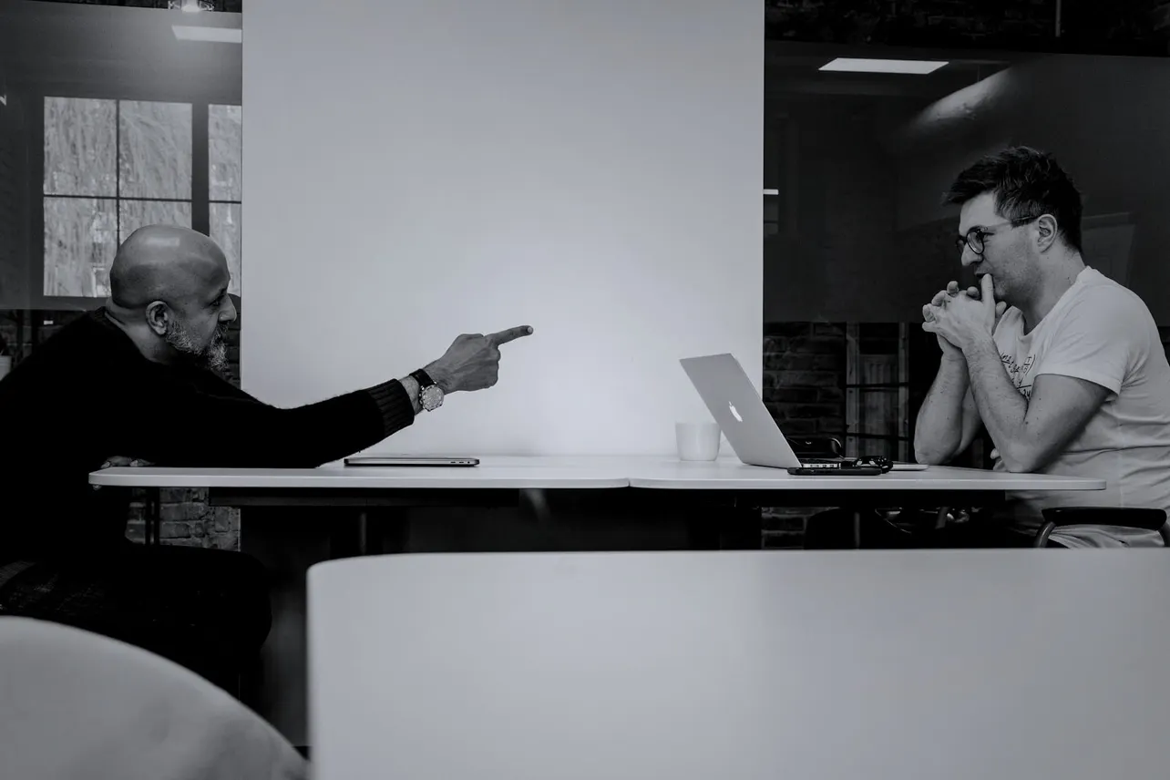 Two men face off in a tense office meeting, one pointing, the other contemplative. A snapshot of modern workplace friction.