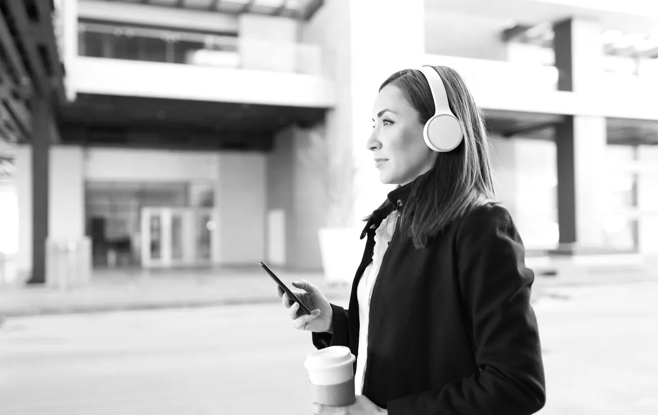 Woman wearing headphones and holding coffee, listening to the EBN Dispatch podcast outside an office building.
