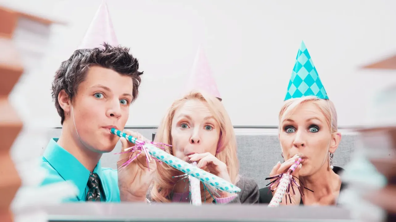Three office colleagues in party hats leaning over a cubicle wall, enthusiastically blowing noisemakers, with blurred stacks of paperwork framing the scene.