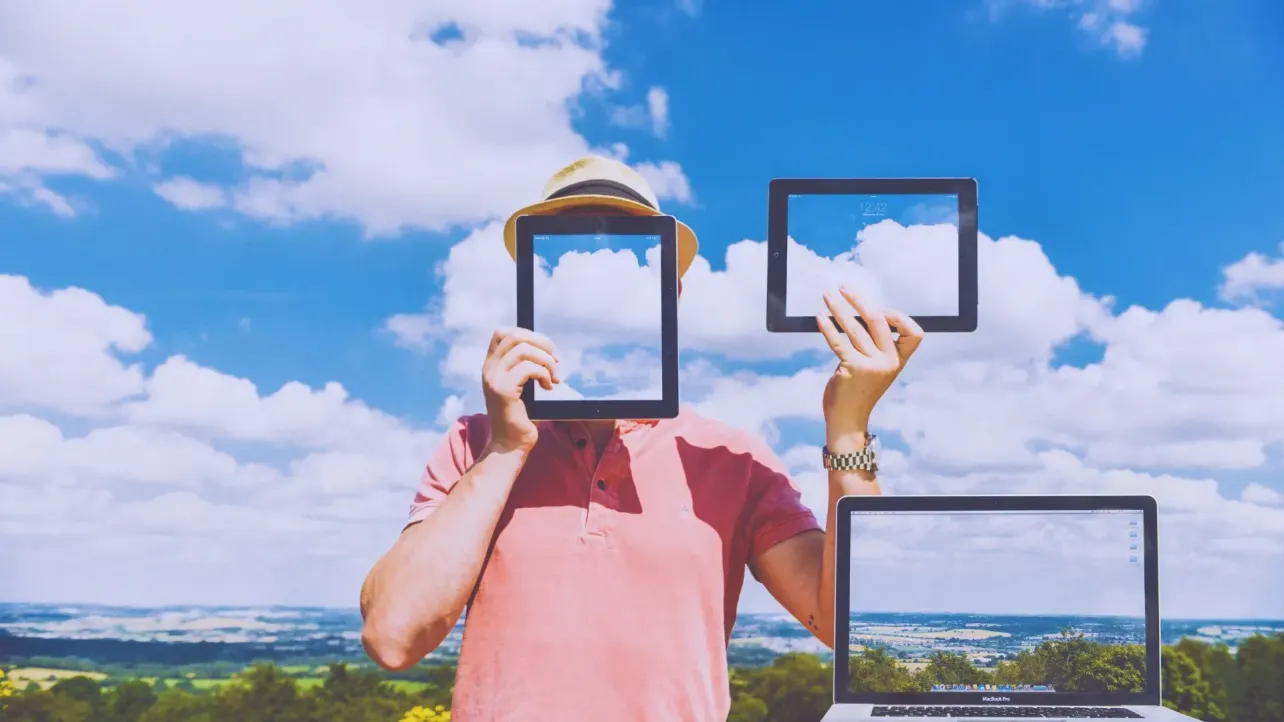 Person in straw hat and pink polo holds two tablets framing clouds; a laptop echoes the view, replacing their face with sky to show how screens blur personal identity.