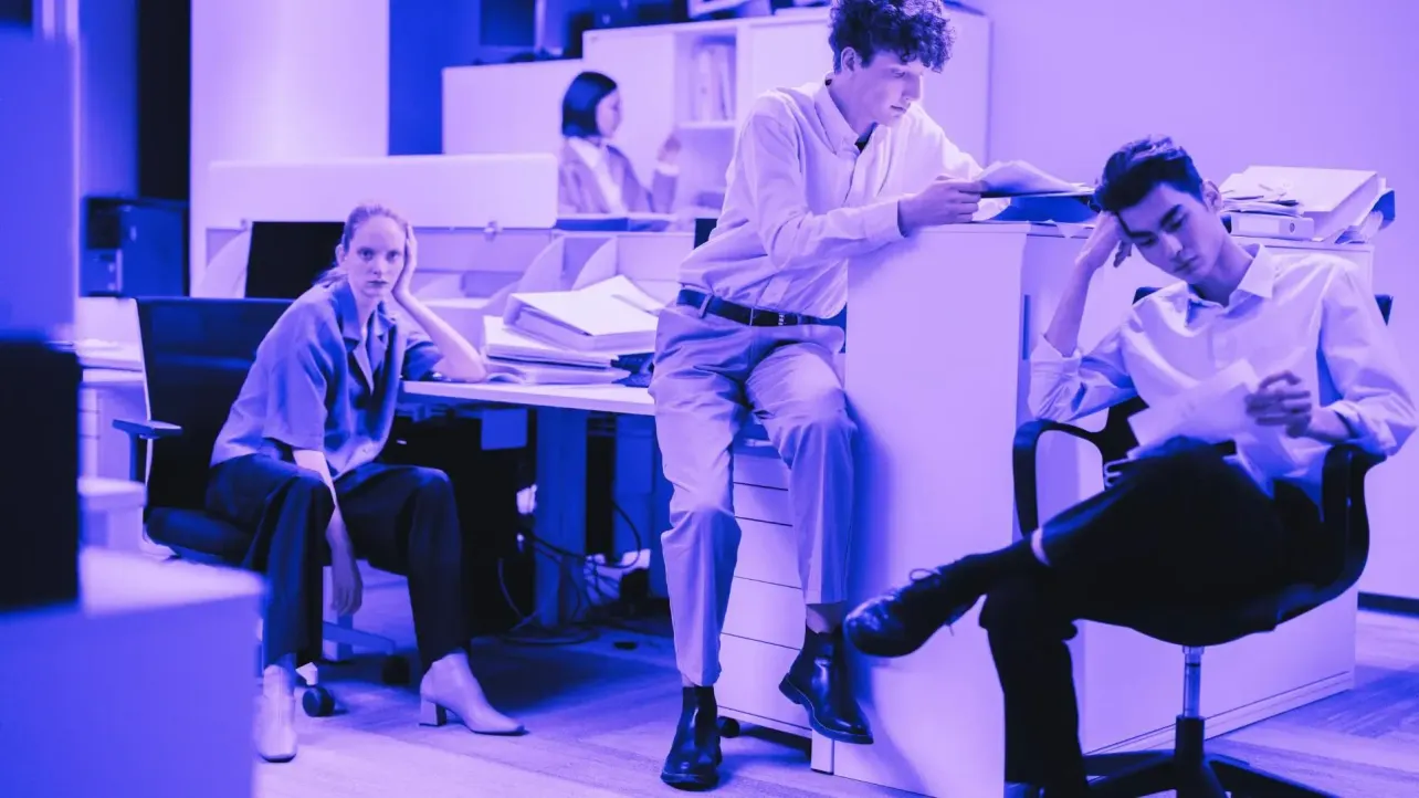 Purple-washed office tableau—four young professionals lounged in weary poses among immaculate desks and scattered papers, underscoring the gap between curated teamwork and real-life morale.