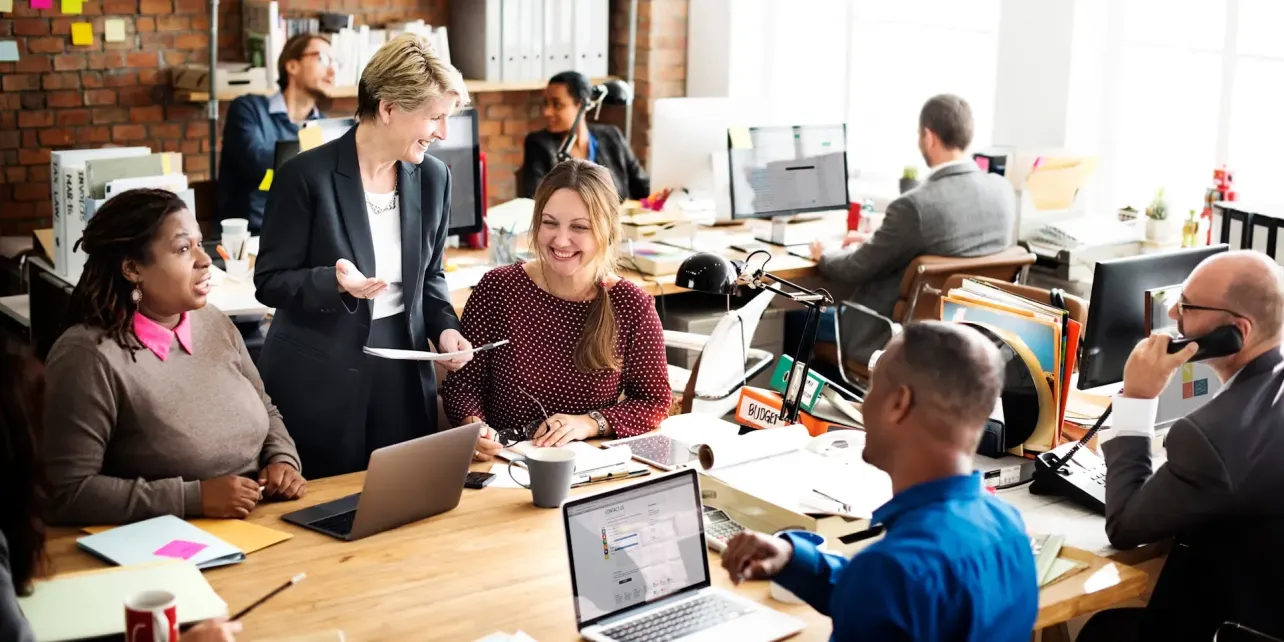 Diverse group of professionals collaborating and laughing around a shared desk in a bright, open-plan office.