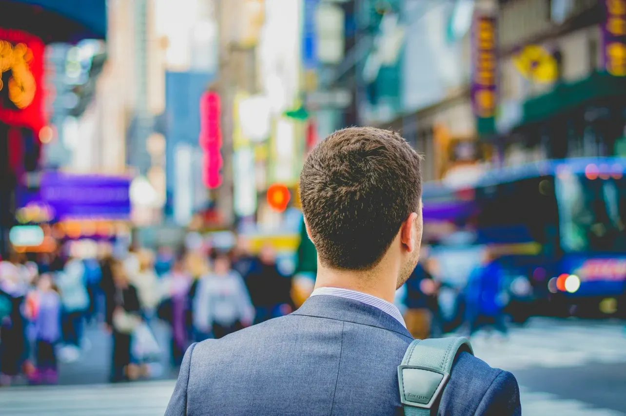 Back view of a business professional, standing in a vibrant city street filled with people and colorful signs, representing career possibilities and workplace culture.