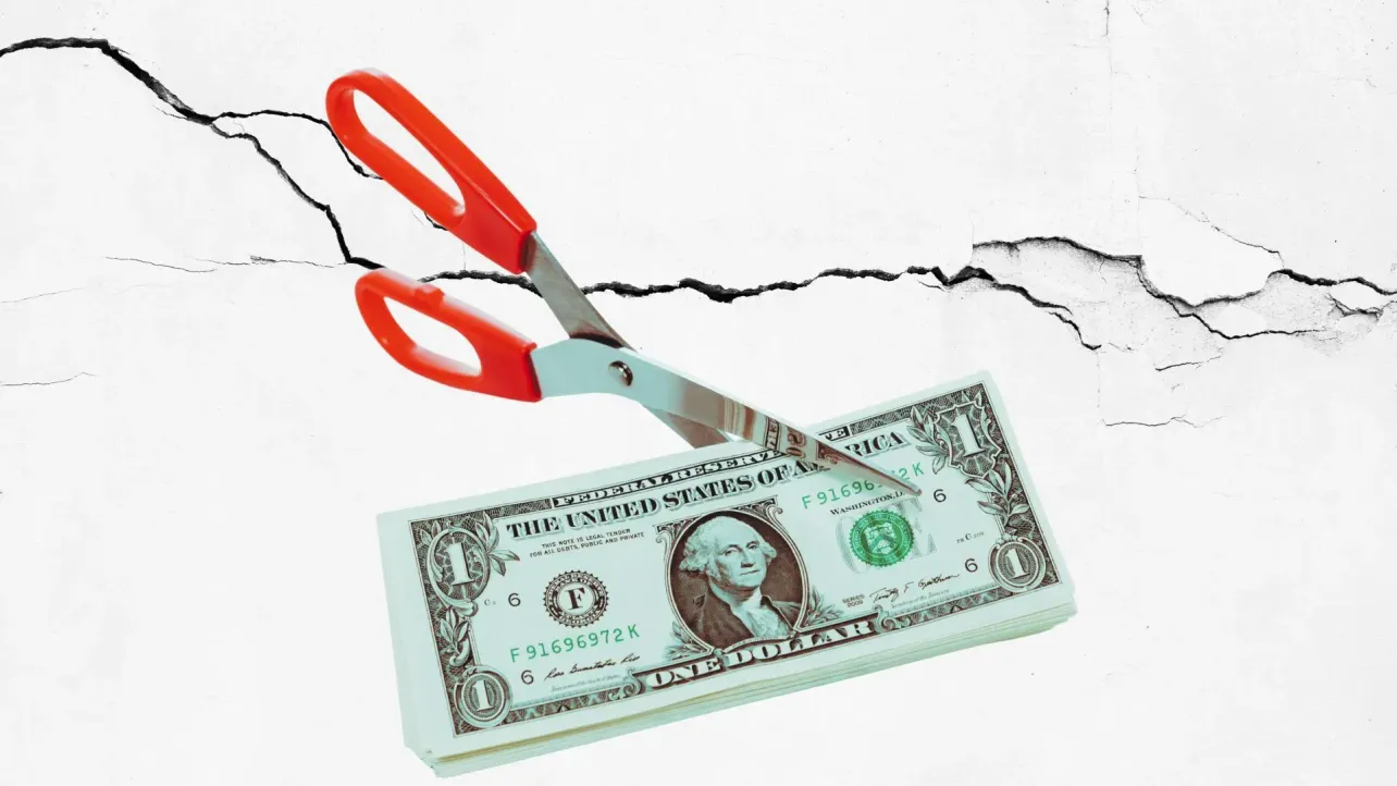 Close-up of scissors slicing through a stack of one-dollar bills against a cracked concrete wall, symbolizing cost cutting that weakens foundations and creates hidden costs.