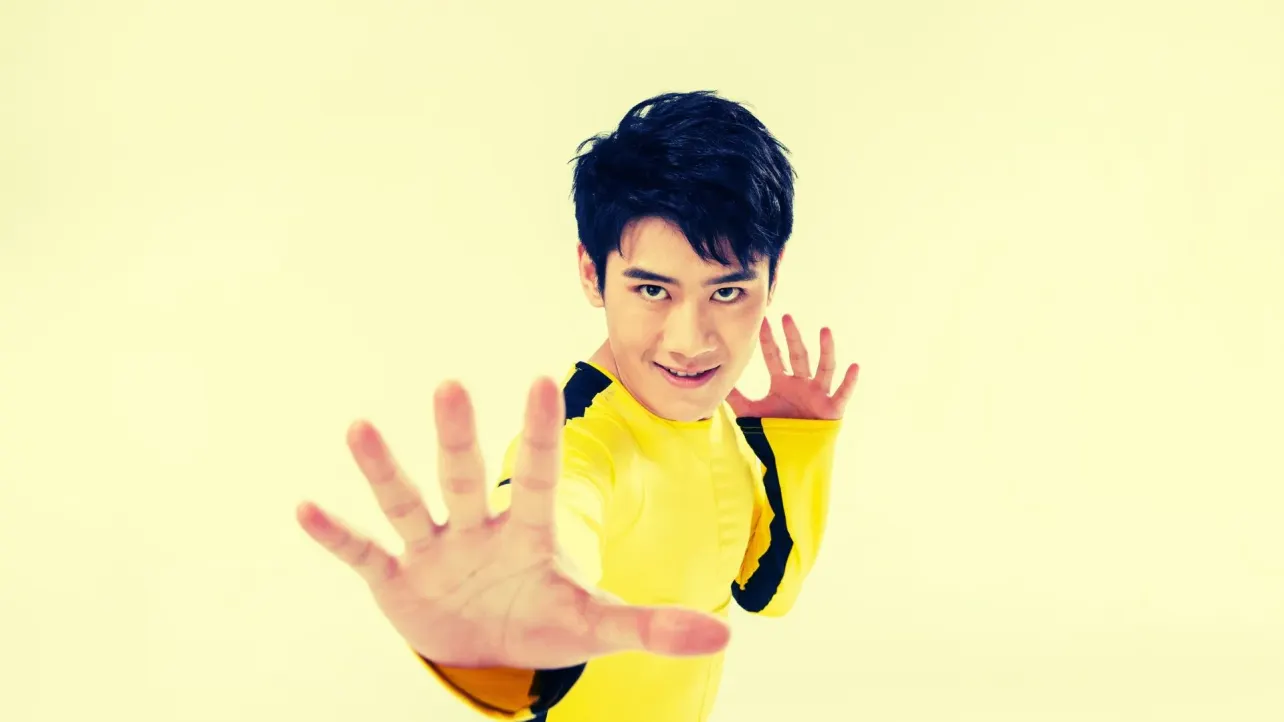 Martial artist in a yellow jumpsuit reaches toward the camera with an open palm in a Bruce Lee style pose against a pale studio background.