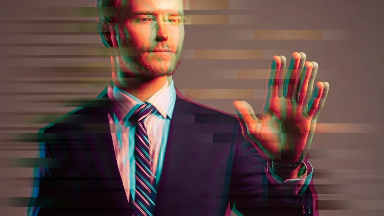 A suited professional holding up a hand in a stop gesture, with a glitch effect across the image, suggesting identity manipulation and digital deception in hiring.