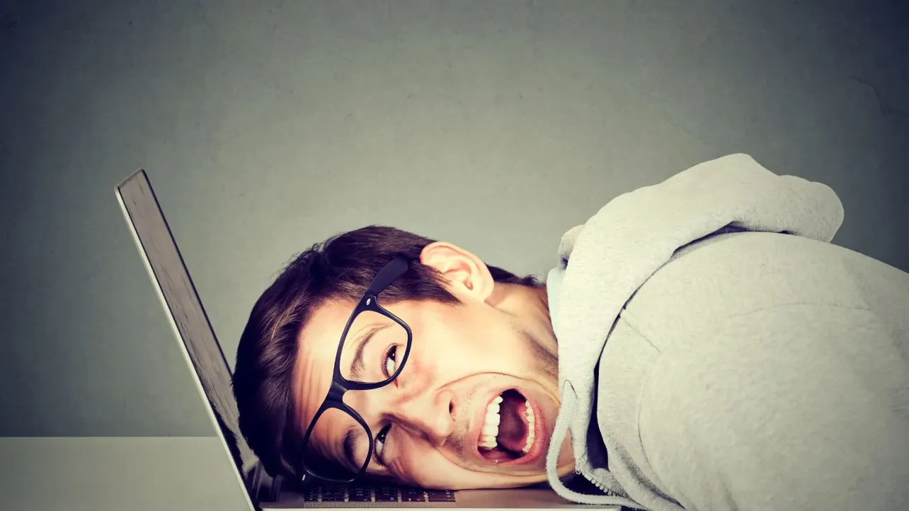 A person in glasses lying face-down on a laptop keyboard with their mouth open in frustration, suggesting burnout and work overload.