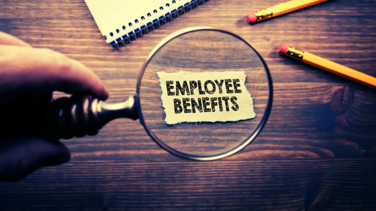 A magnifying glass highlighting the words “Employee Benefits” on a desk, suggesting scrutiny of workplace perks and policy changes.