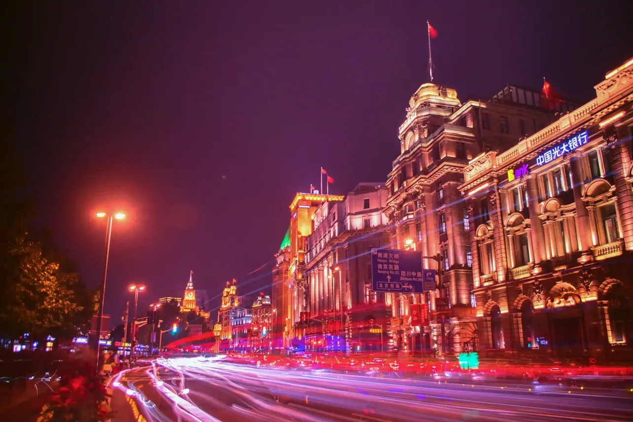 Night view of Shanghai with illuminated historic buildings and long-exposure traffic trails, reflecting the speed and pressure of business in China.
