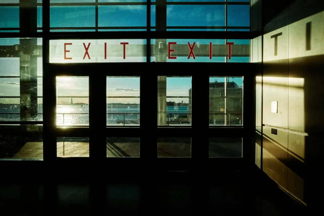 Red exit signs above glass doors, light beyond, shadows in the foreground.