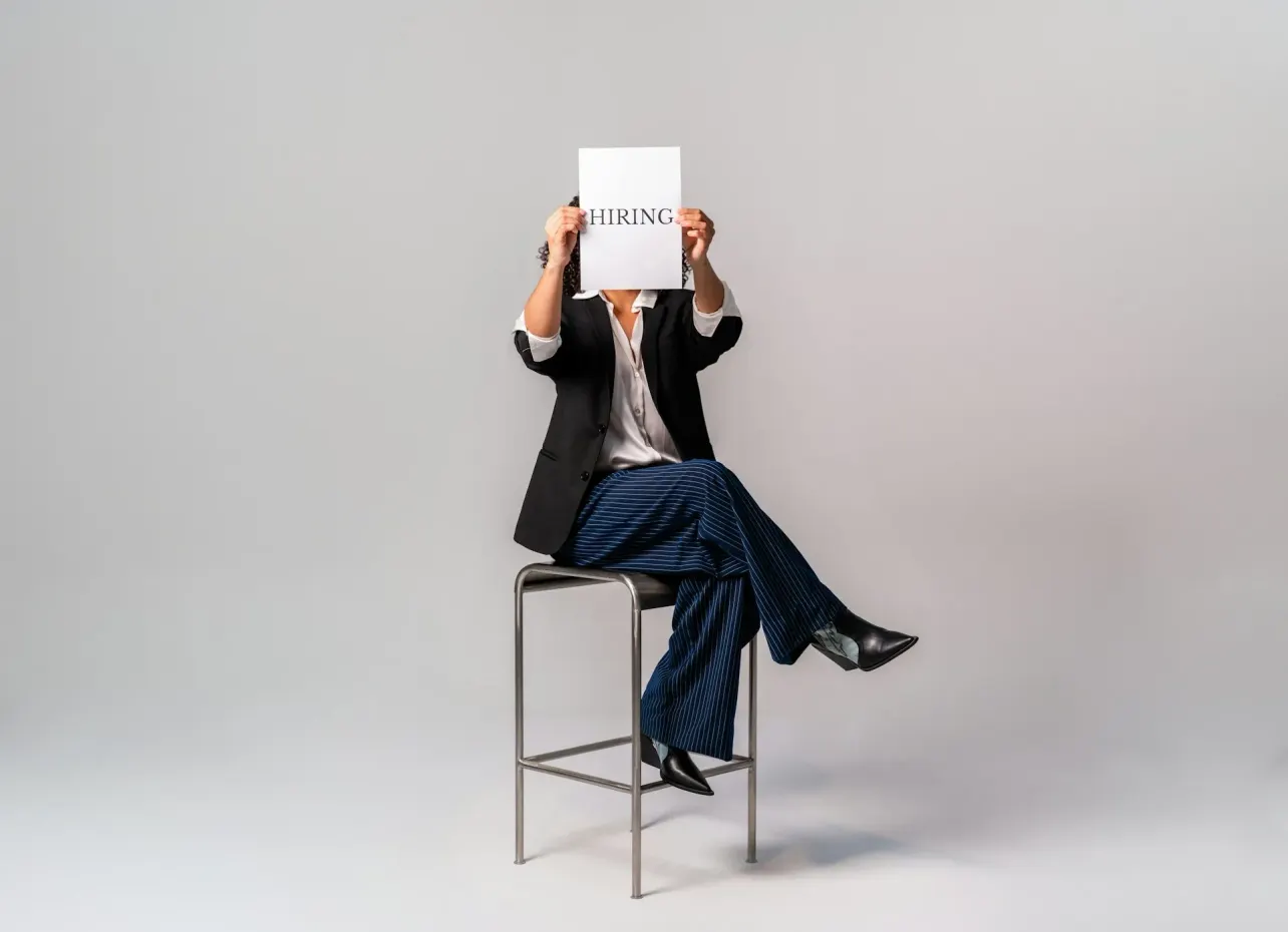 A professional seated on a stool holds a plain white sign reading "HIRING", representing the gap between recruitment signalling and the deeper strategic discipline of employer branding.