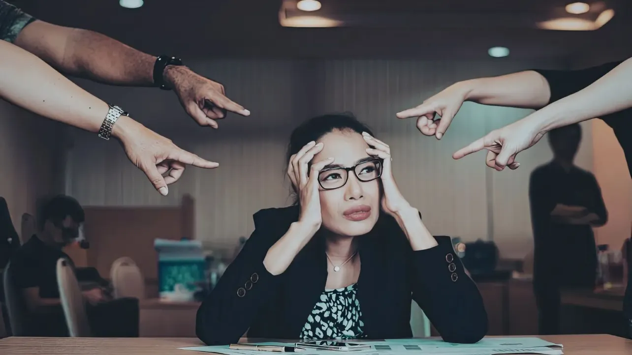 An overwhelmed HR professional holding their head while multiple hands point at them, suggesting workplace pressure, blame, and organisational stress.
