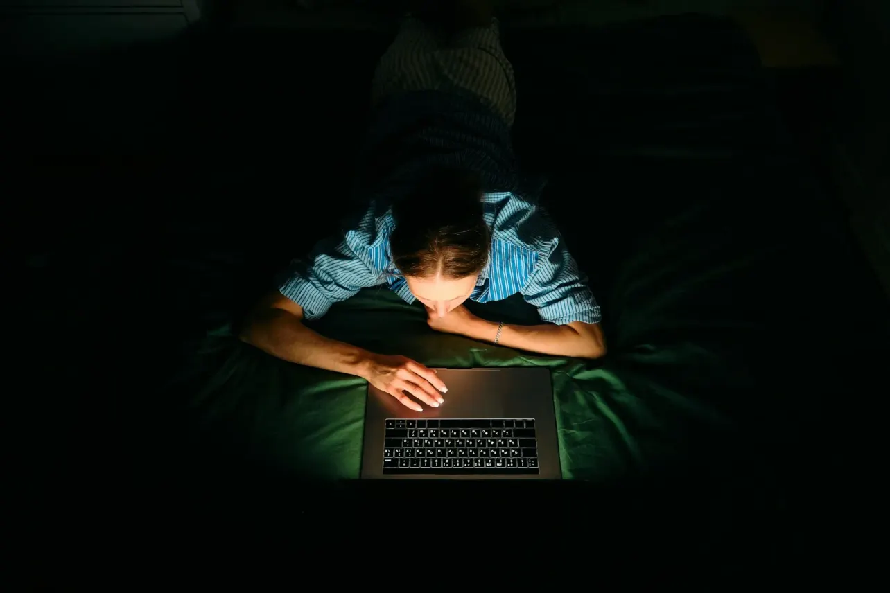 Overhead view of a woman lying in a dark room, face and hands lit only by her laptop screen as she researches late at night.