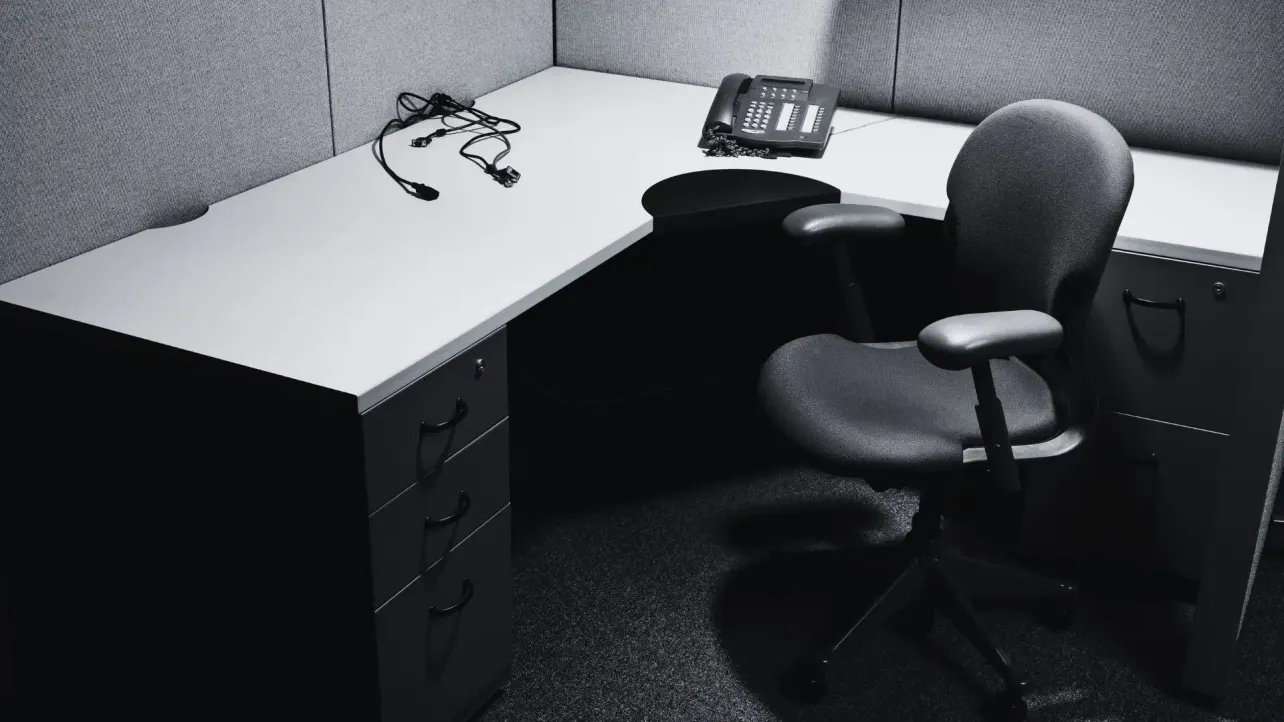 An empty office cubicle in black and white, chair pushed in and a desk phone unplugged with its cables coiled loose, symbolising a role removed by decision rather than by a person.