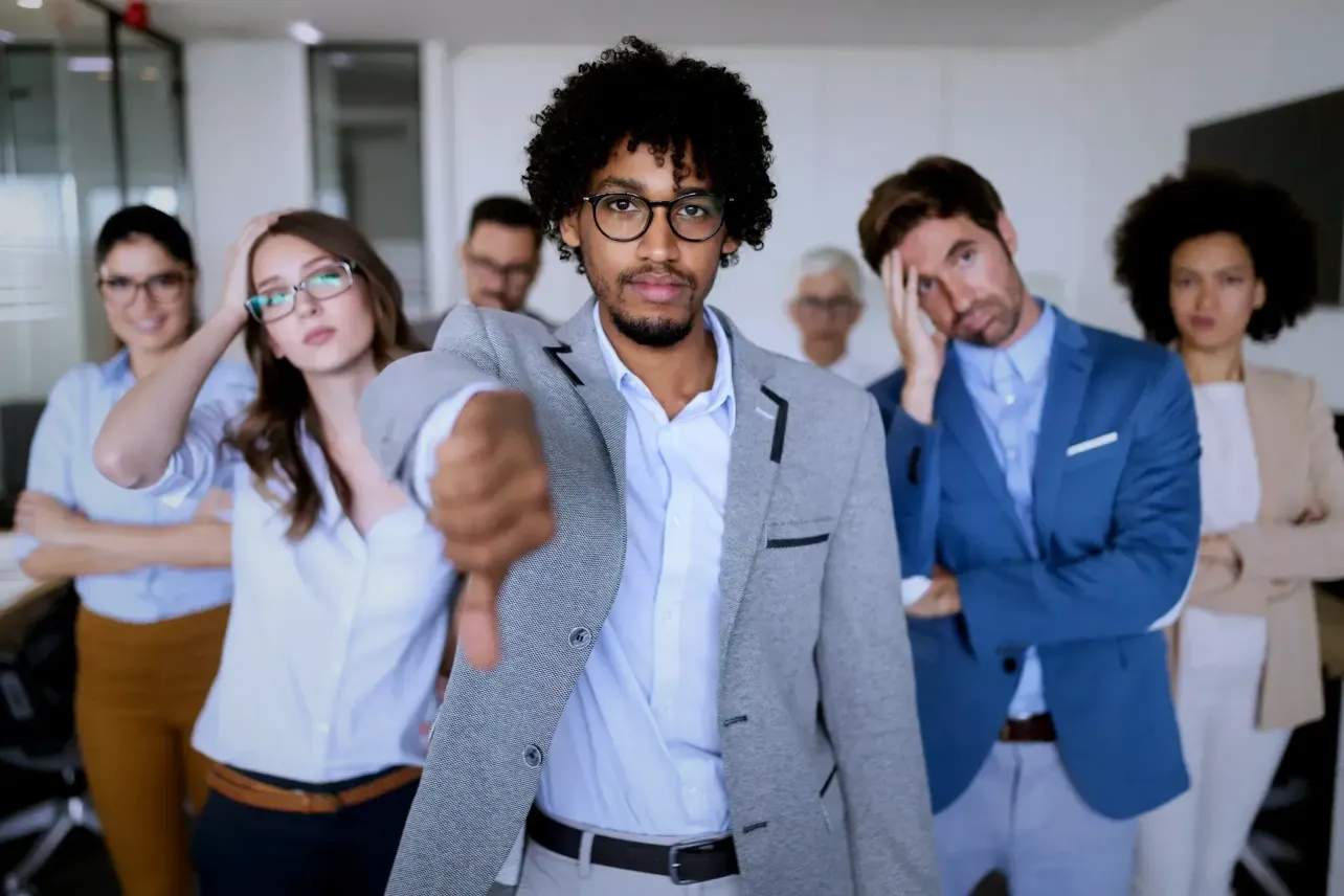A frustrated office team stands behind a man giving a thumbs down, illustrating the gap between employer branding claims and employee reality.