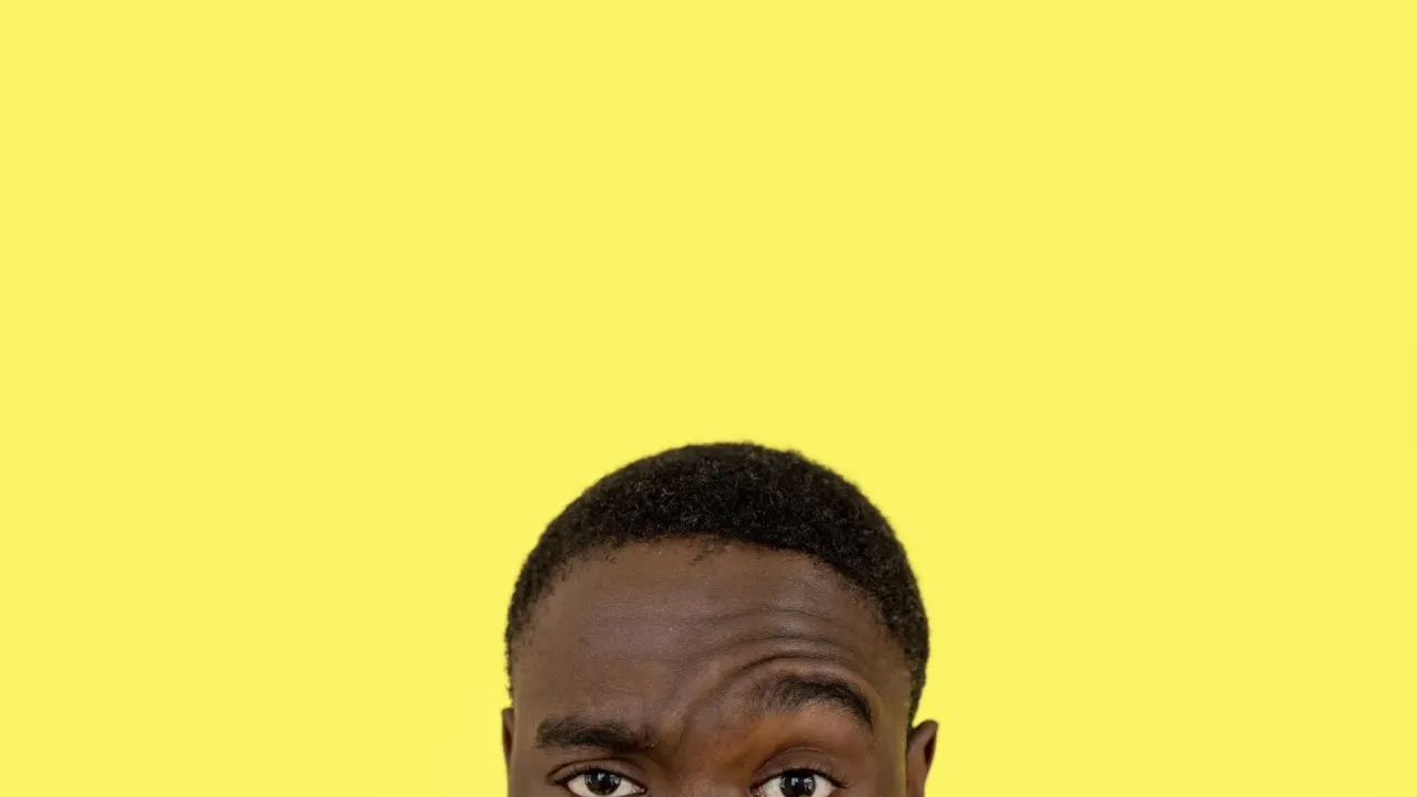Cropped close on a man's raised eyebrow and narrowed eye, a sceptical look, against a flat yellow background.