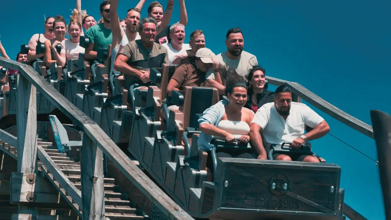 A full car of rollercoaster riders cresting the top of a hill, reacting with a mix of excitement, fear and hands raised.