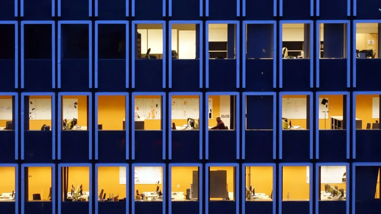 A grid of lit, mostly empty office rooms behind a blue facade at night, with one person working at a desk