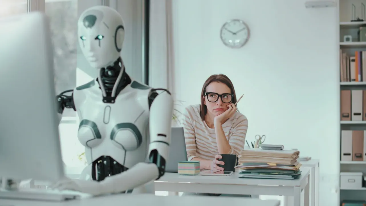 A humanoid robot working at an office keyboard while a woman sits nearby watching, unimpressed, with a coffee mug.