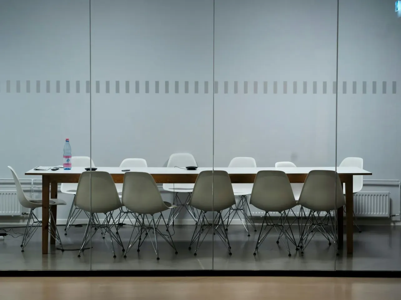 Empty modern conference room with a long meeting table and white chairs behind glass walls.