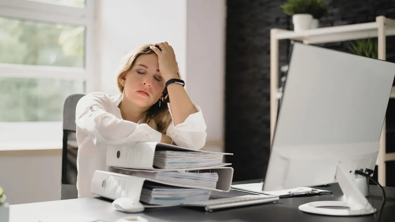 Exhausted office worker slumped over binders at her desk, illustrating this week's Paper Cut on layoffs and rising workloads.