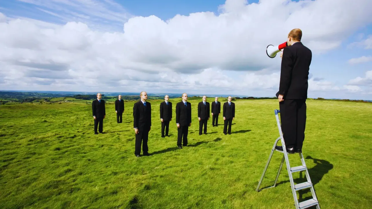 Man on a stepladder shouts through a megaphone at eight identical men in suits standing in a field