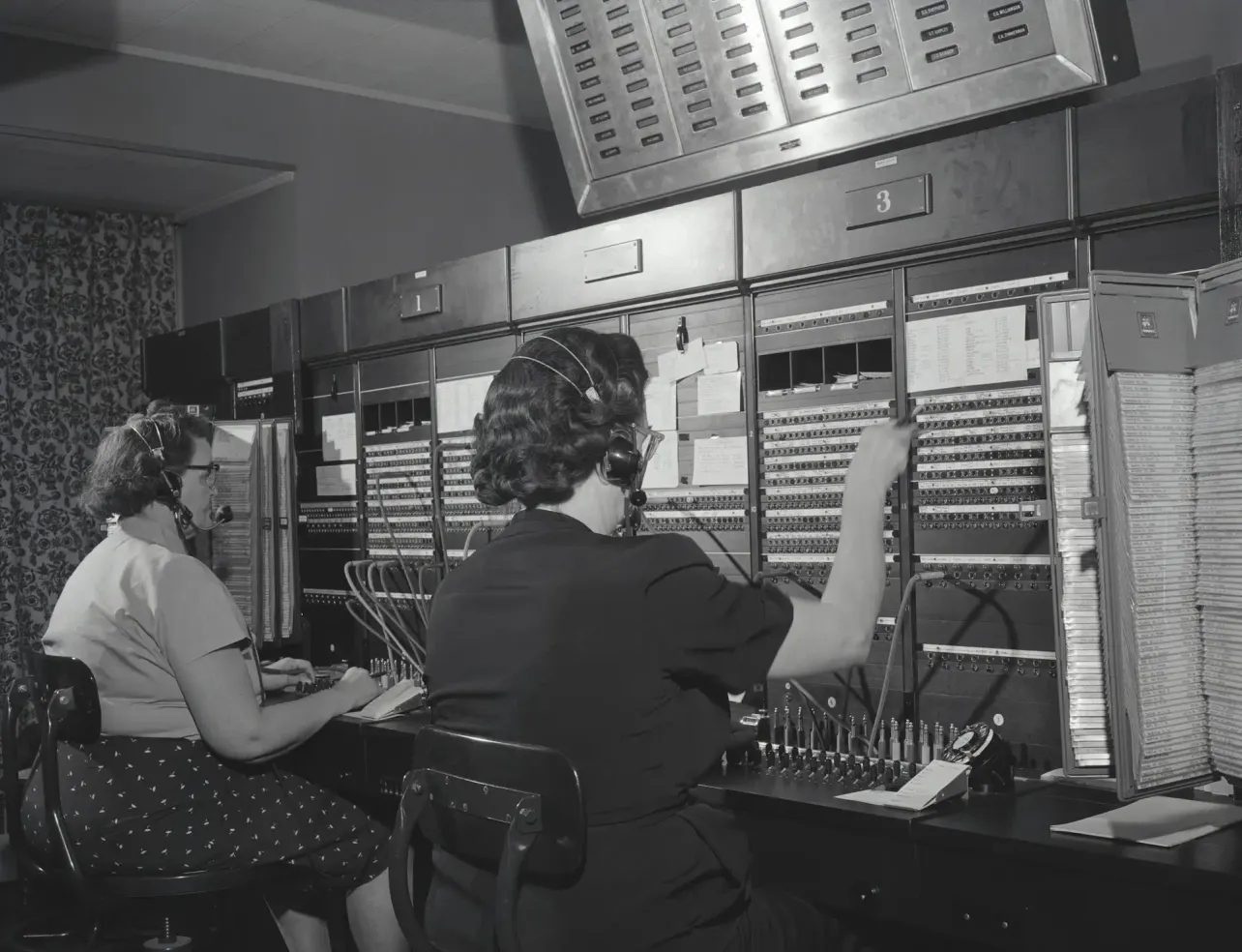 Two telephone operators wearing headsets and connecting calls at a large manual switchboard in a black-and-white photograph.