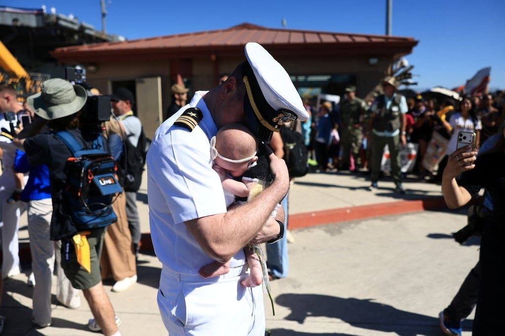 An American sailor in uniform embraces his newborn daughter just as he has disembarked from his ship. His face is buried in the crook of her neck.
