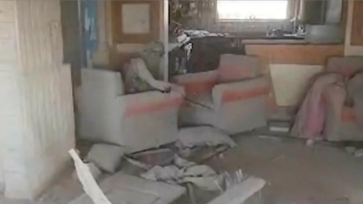Man sitting in chair in a room damaged by war, his face and head covered in a green scarf looks into camera