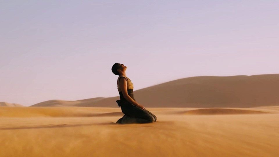 A woman is kneeling and wailing at the sky; the background is a bleak, open desert