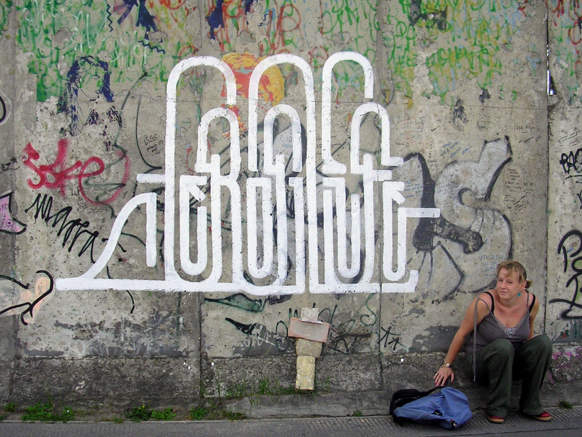 A young blonde woman sits against a grey section of the Berlin Wall with bright white graffiti art in the center.