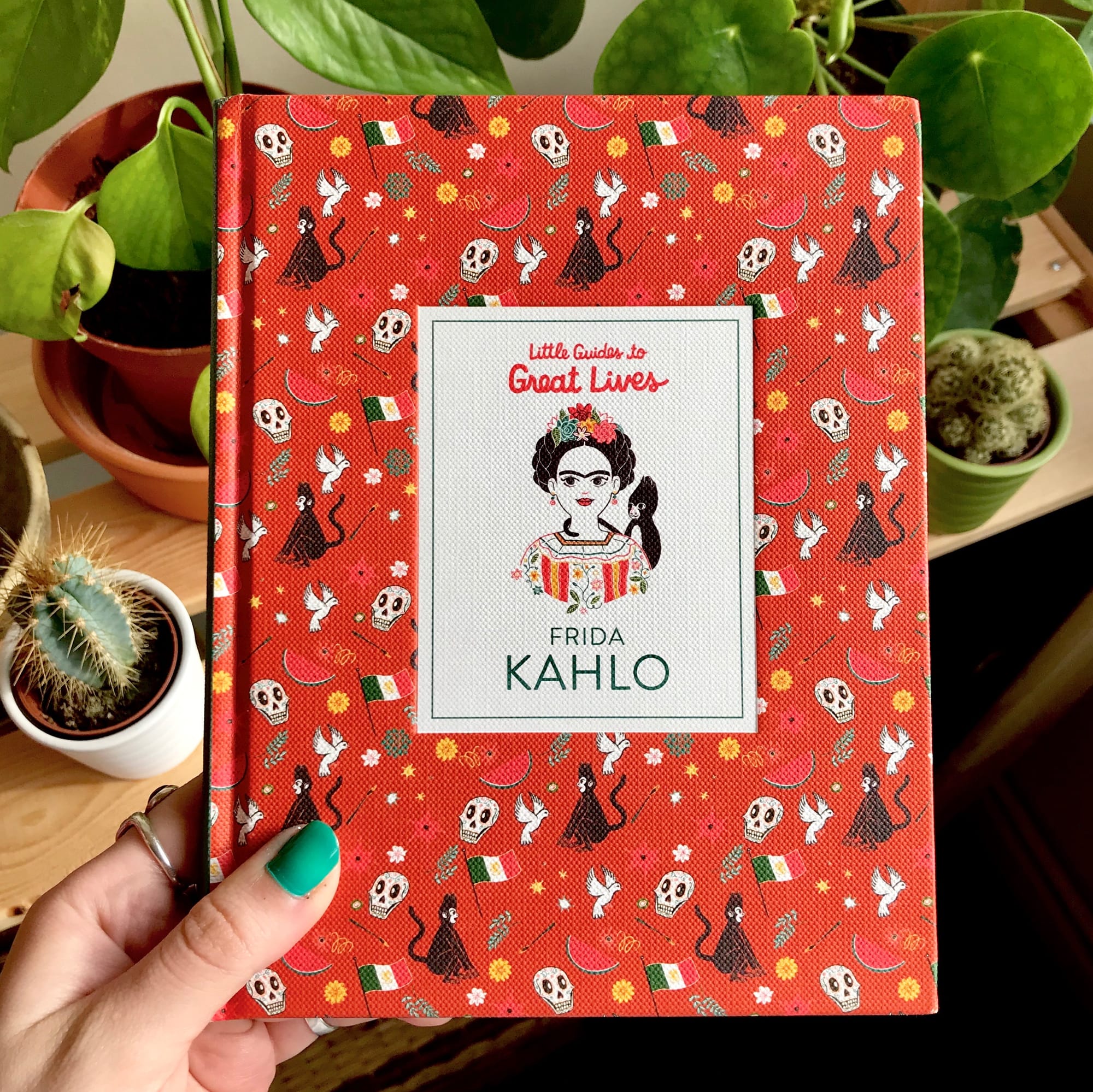 Hand with painted green nails holding book cover against plant foliage