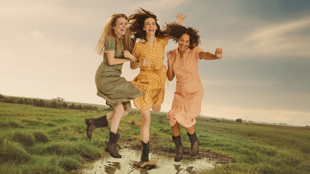 Three young women wearing knee-length light summer dresses and boots jump into a muddy puddle in the middle of a grassy field.