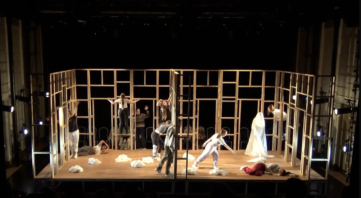A wooden structure surrounds the stage. A group of performers can be seen on stage - some are climbing the structure, some are lying on the floor and one is stood up covered in a white sheet.
