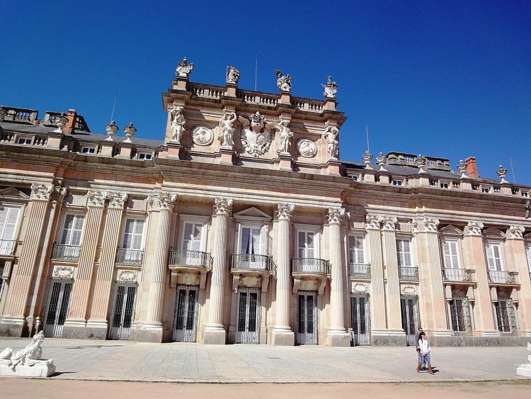 The sunlit façade of the Royal Palace of La Granja de San Ildefonso beneath a clear blue sky.