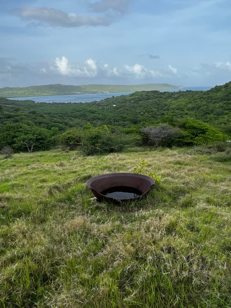 The image is a metal Copper used in colonial times, in a green grassy area with a view of Chirstian Cove and Willoughby Bay, Antigua
