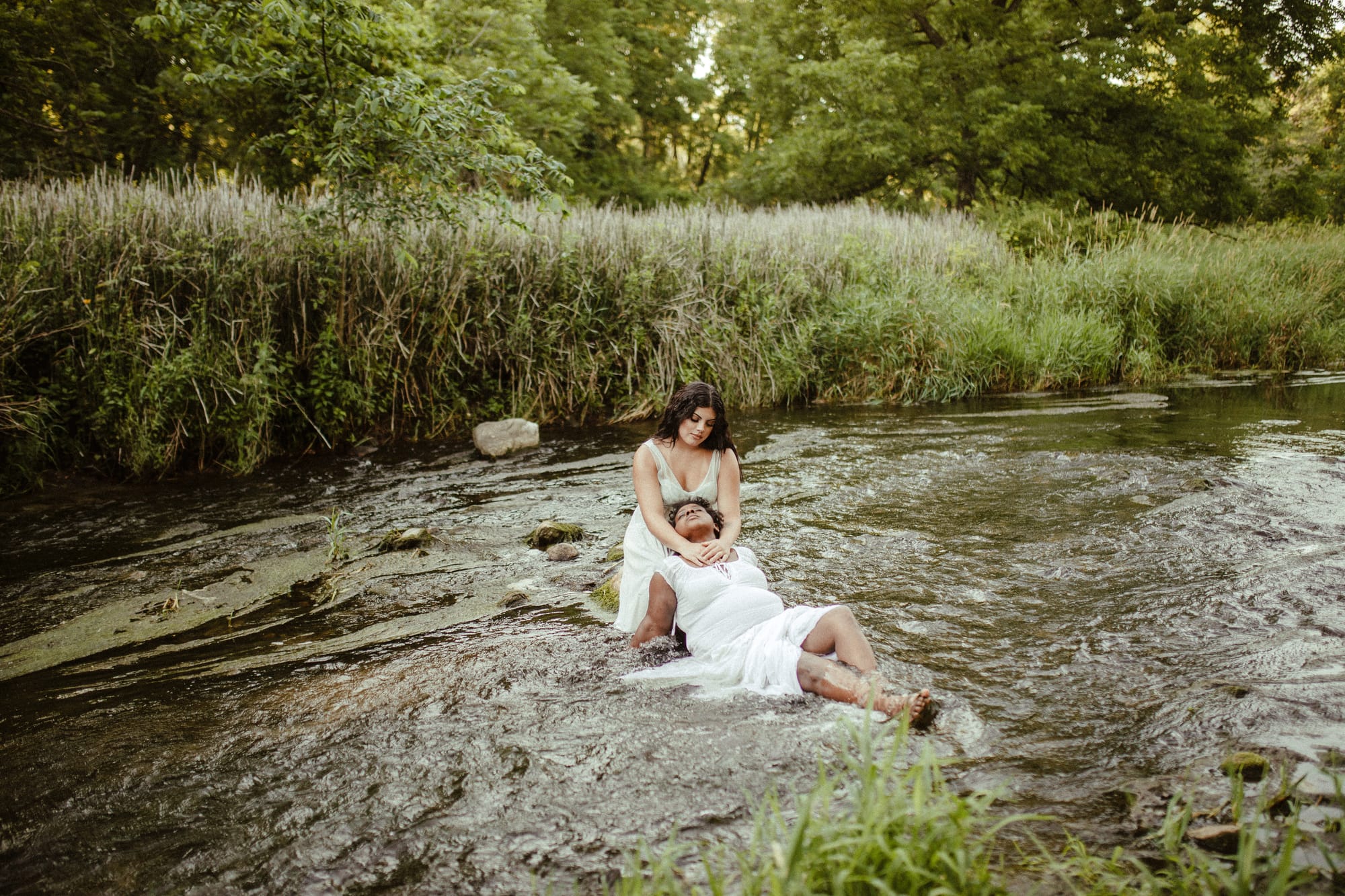 Two women in white, in a creek banked with reeds, one leaning back on the other's knees, the other's hand over the resting woman's heart.
