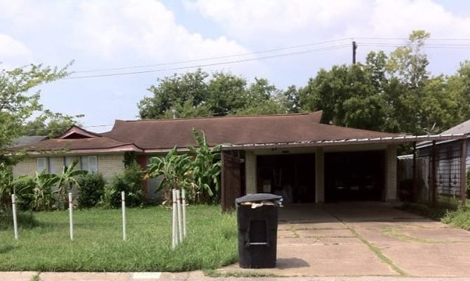 Photo of a one-storey house with two-car carport, palm and live oak trees, and a white pole fence and garbage can awaiting collection..