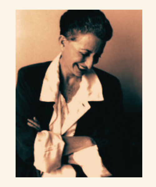 Sepia toned portrait of Cixous with short gray hair, smiling and look down, wear an oversized black suit and white blouse.