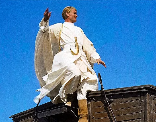 A gif of Lawrence, clad all in white, stepping down from a train and raising his hand 